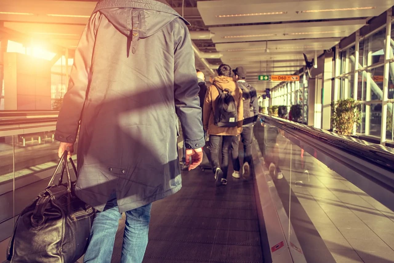 Një udhëtar ecën nëpër një korridor aeroporti të ndriçuar nga dielli me valixhe, drejt pushimeve.
